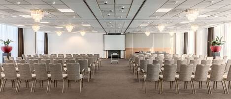 Un salón de conferencias con filas de sillas, una pantalla de proyector y una zona de escenario.