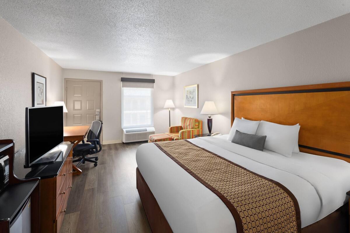 A hotel room with a large bed, a desk with a flat-screen TV, a chair, a lamp, and a window with blinds.