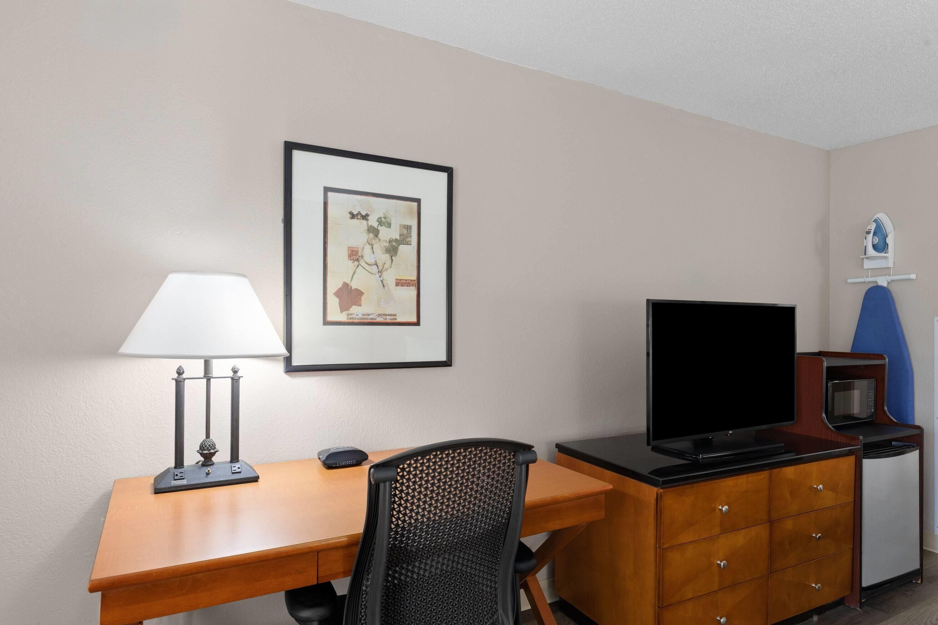 Habitación de hotel con un escritorio de madera, una silla de oficina negra, un televisor de pantalla plana, un microondas y una obra de arte enmarcada en la pared.