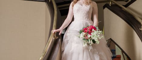 Una persona con vestido de novia bajando una escalera con barandillas ornamentadas y alfombra estampada.