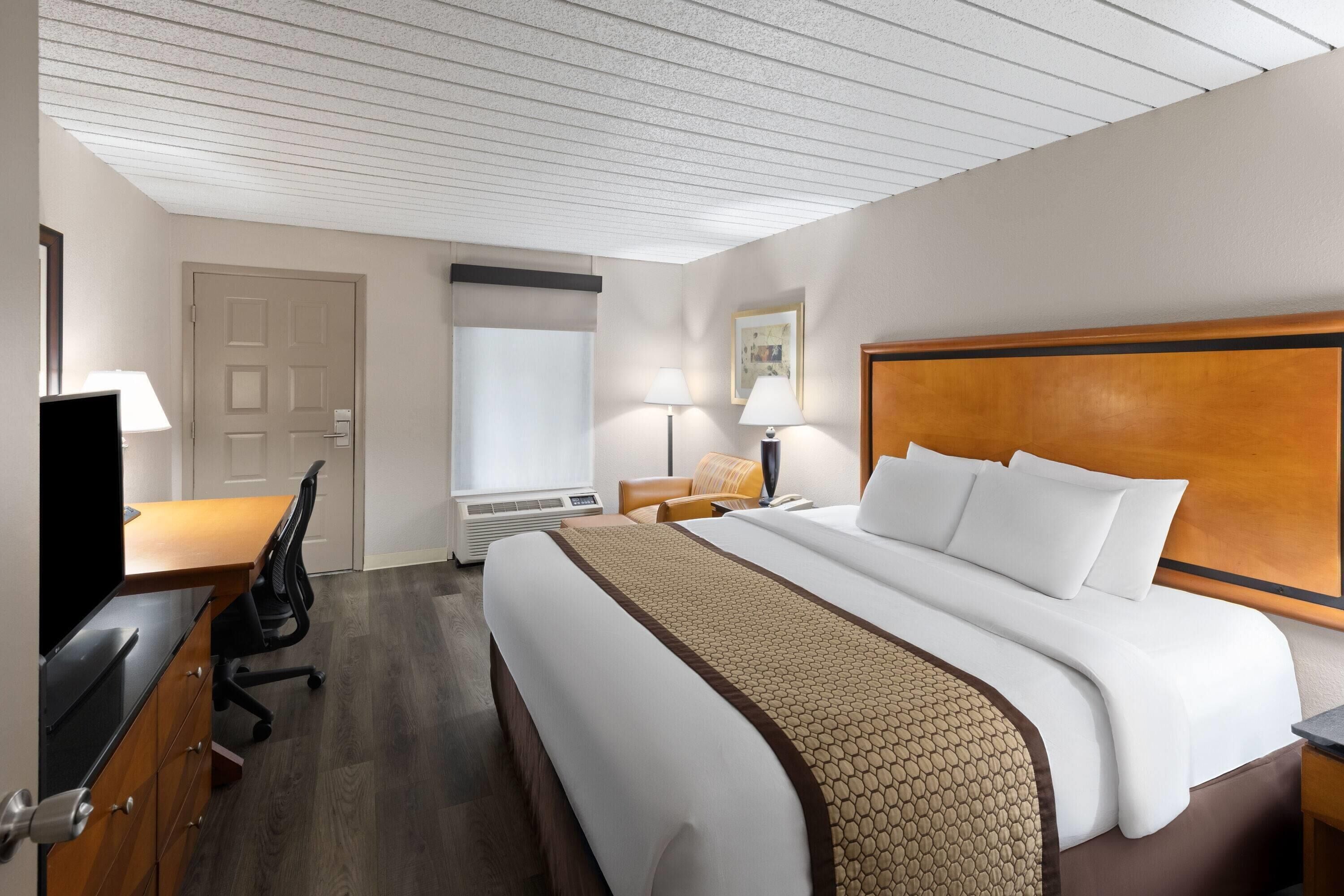 Quarto de hotel com uma cama grande, uma escrivaninha com cadeira, uma televisão e uma janela com cortinas.