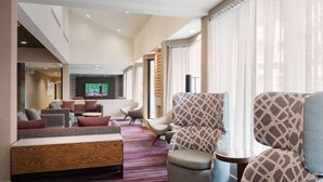 Un moderno vestíbulo de hotel con un área de descanso que incluye sofás y sillones, un televisor de pantalla plana y ventanas amplias con cortinas translúcidas.