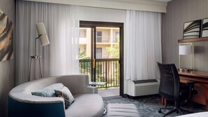 Habitación de hotel con sofá curvo, escritorio, silla, lámpara de pie y vistas al balcón exterior.