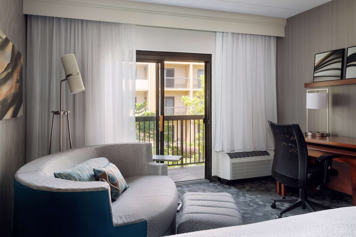 A hotel room with a curved sofa, a desk, a chair, a standing lamp, and a view of the outside balcony.