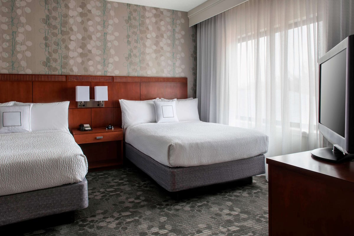 A hotel room with two beds, a wooden headboard, a nightstand with lamps, a television, and a large window with sheer curtains.