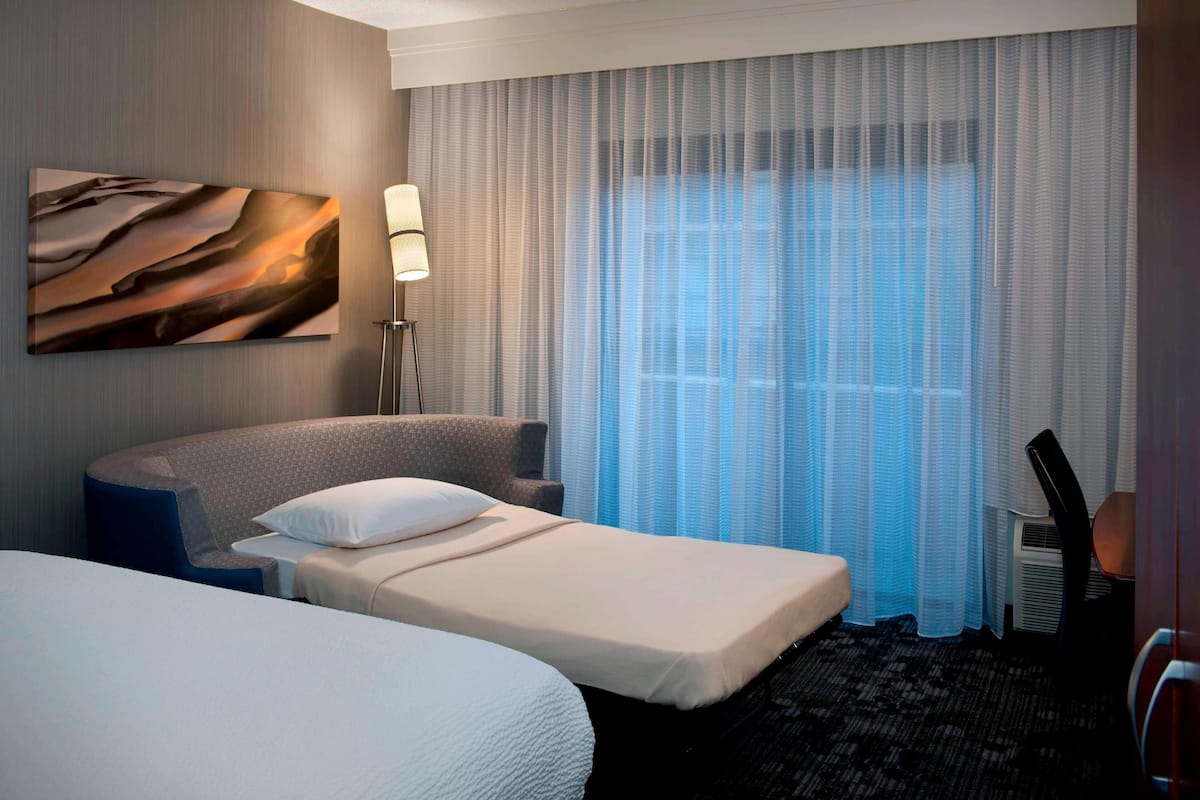 A hotel room with two beds, a large window with sheer curtains, a wall-mounted lamp, and a painting.