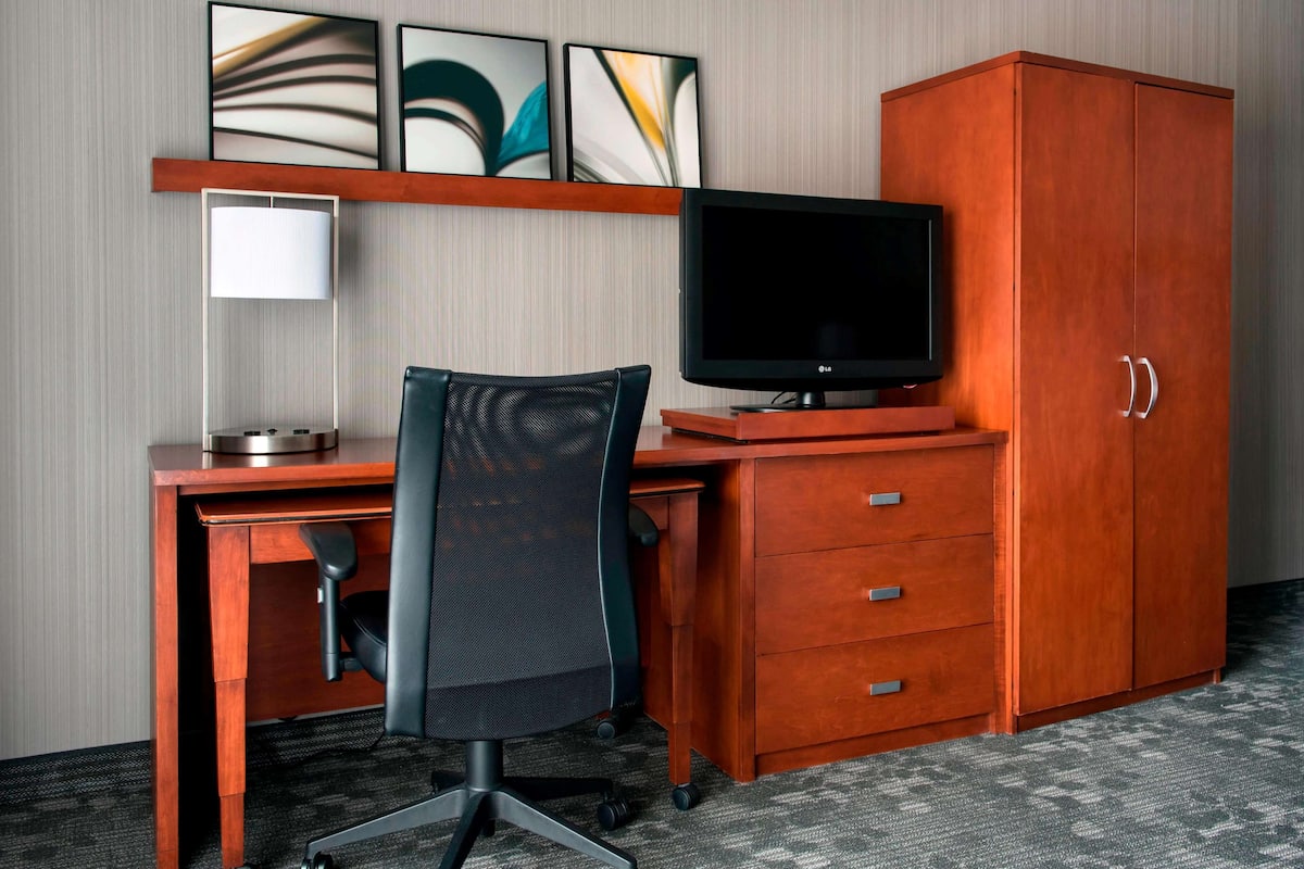 A hotel room with a wooden desk, a black office chair, a flat-screen TV, a lamp, and a wardrobe.