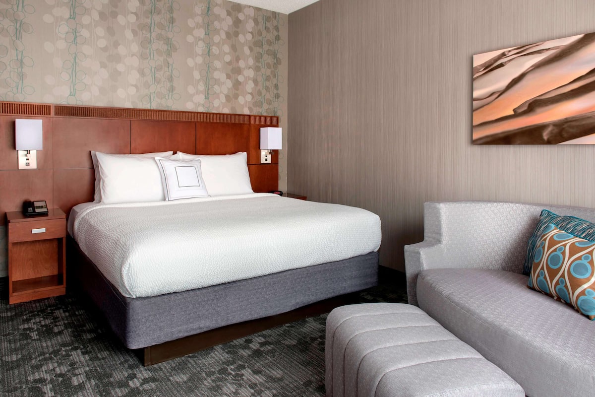 A hotel room with a large bed, a nightstand, a bedside table, a sofa, and a wall-mounted artwork.