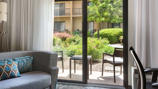 Una habitación de hotel moderna con un sofá gris, una almohada estampada, una lámpara de pie y un ventanal corredero que da a un patio exterior con una mesa, sillas y vistas a un jardín.
