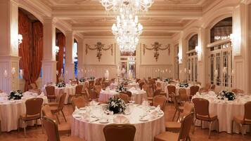 Un vaste hall de banquet, orné de lustres somptueux, avec des tables rondes dressées pour le repas et de magnifiques arrangements floraux en guise de décoration centrale.