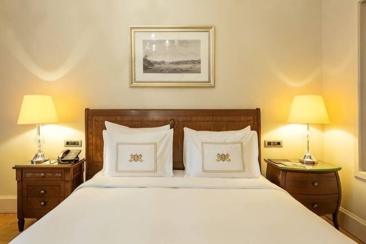 A neatly made bed with two pillows, a wooden headboard, a bedside table with a lamp, and a framed picture on the wall.