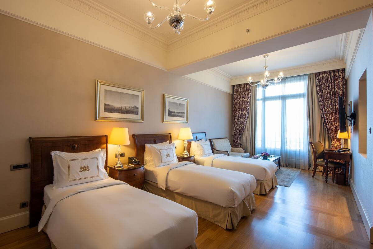 A hotel room with three beds, a chandelier, and a large window with curtains.