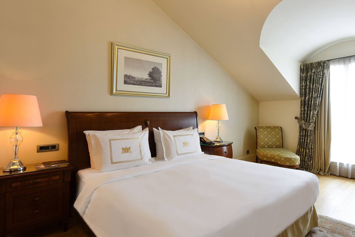 A hotel room with a large bed, two bedside lamps, a framed picture on the wall, a chair, and a window with curtains.