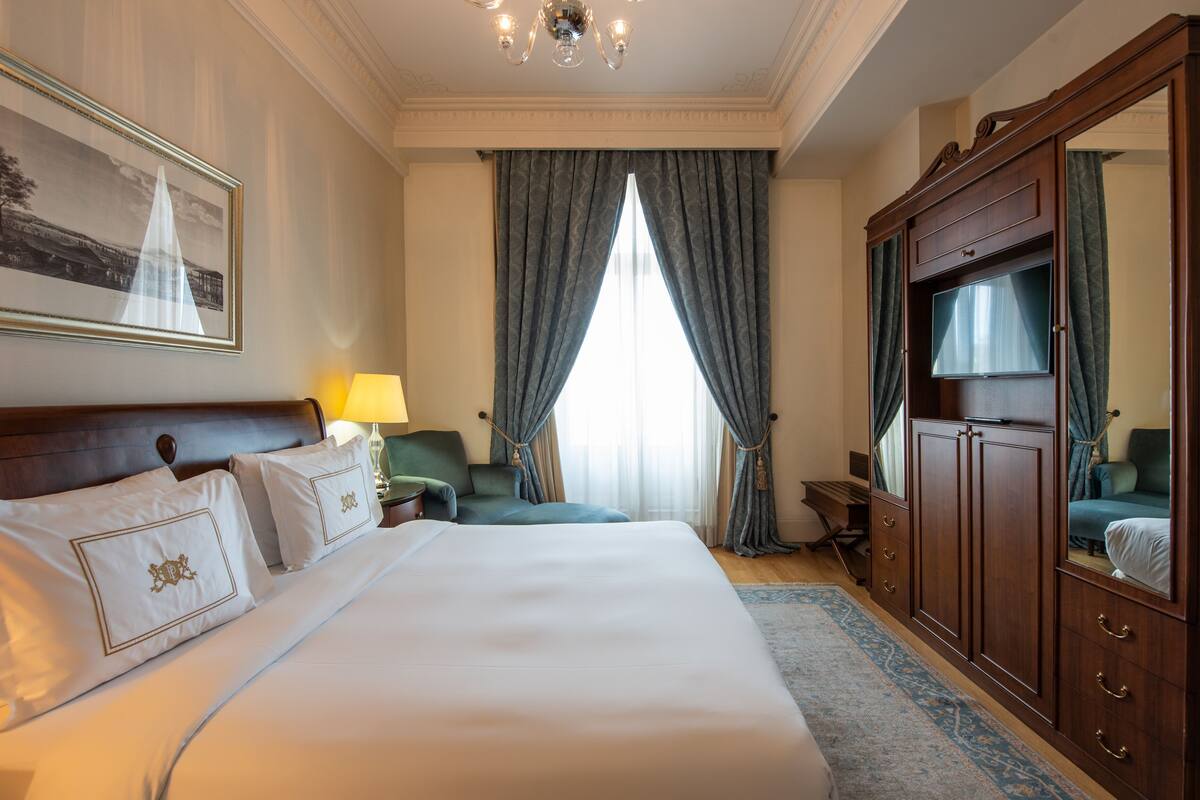 A hotel room with a large bed, a desk, a chair, a wardrobe, and a television.