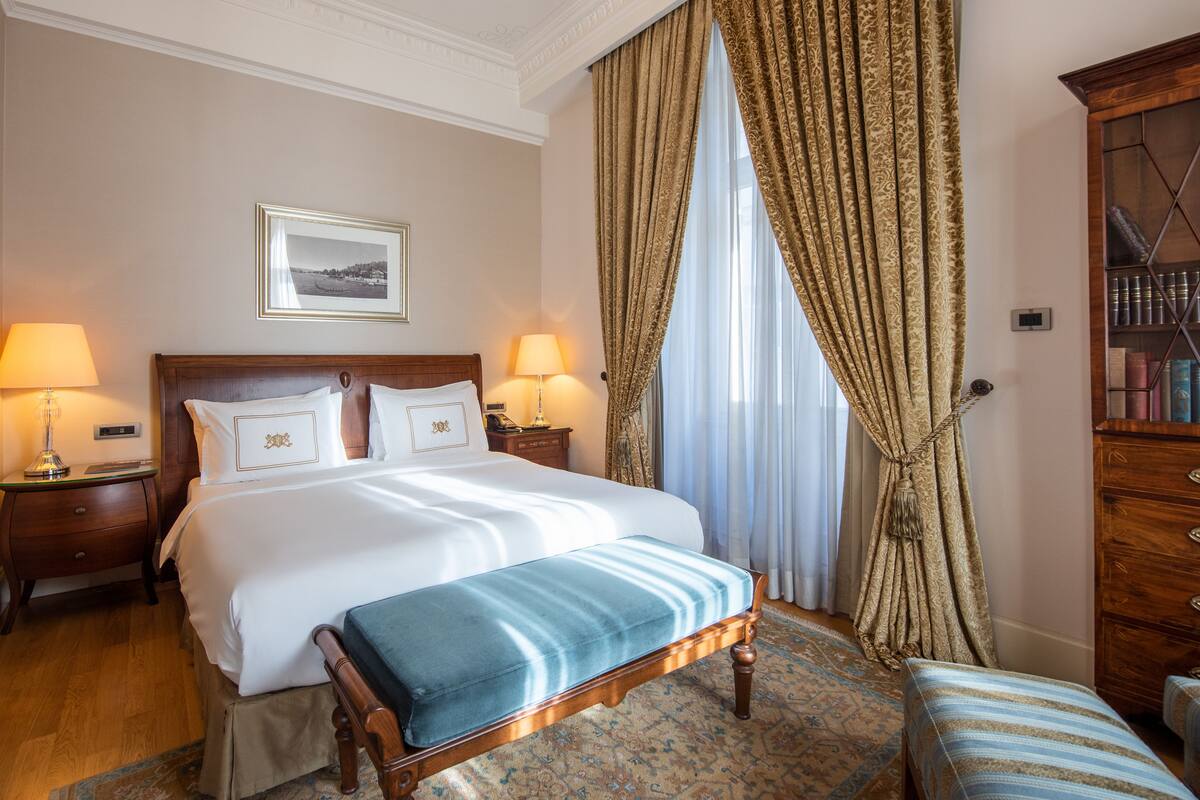 A hotel room with a large bed, two bedside tables with lamps, a wooden bench, a patterned rug, and a bookshelf.