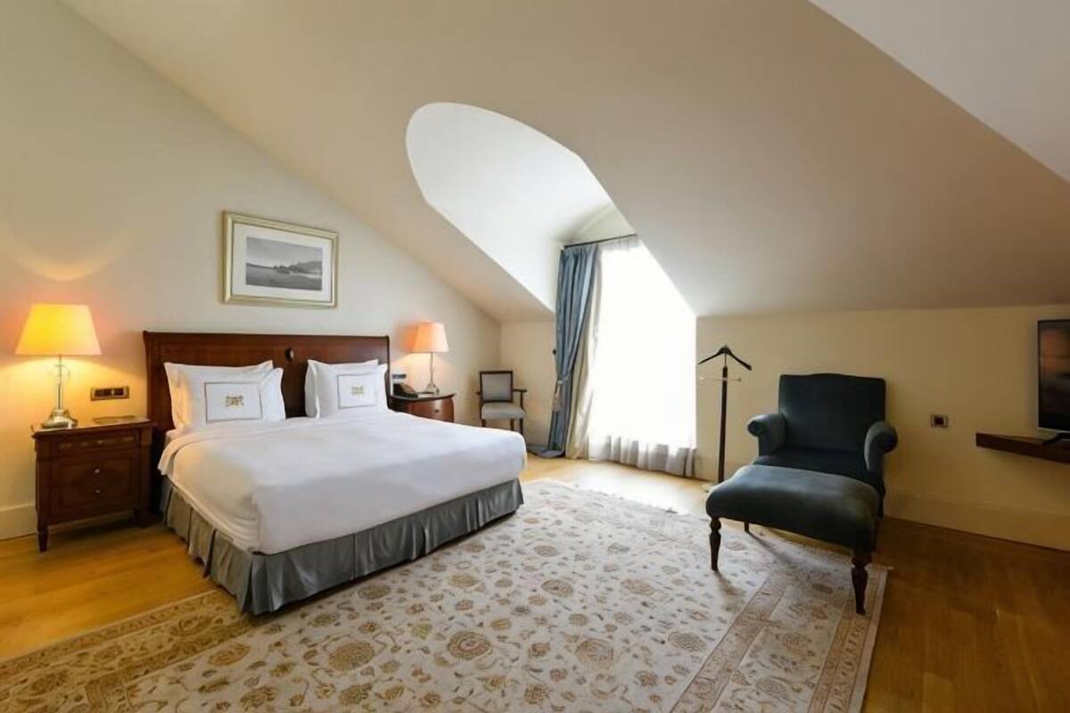 A bedroom with a bed, nightstands, a chair, a desk, and a large window.