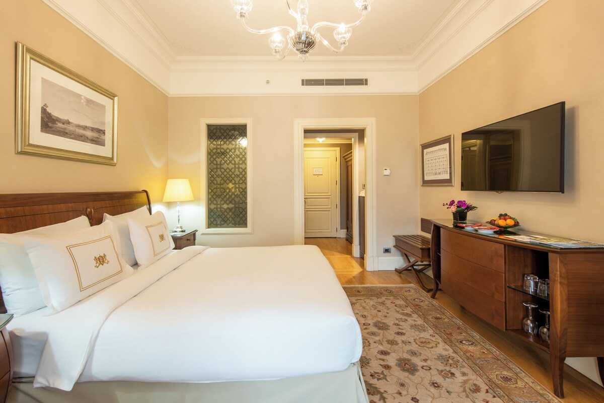 A hotel room with a large bed, a desk with a flat-screen TV, a dresser, and a chandelier.