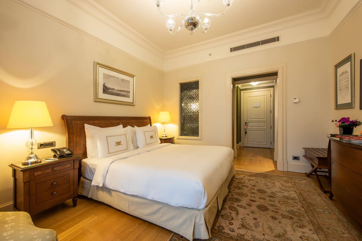 A hotel room with a large bed, two bedside lamps, a nightstand, a wooden dresser, and a chandelier.