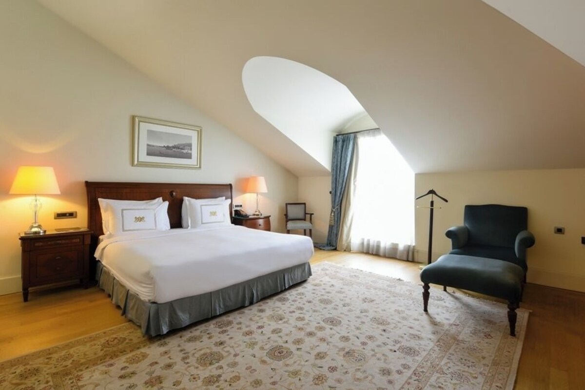 A bedroom with a large bed, two bedside tables with lamps, a chair, a television on a stand, and a framed picture on the wall.