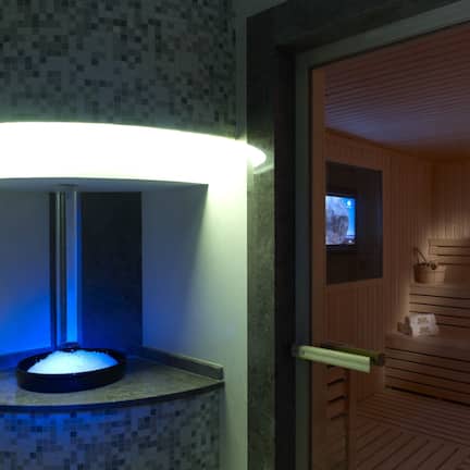 A modern bathroom with a mosaic tile wall, a blue-lit sink, and a wooden sauna visible through an open door.