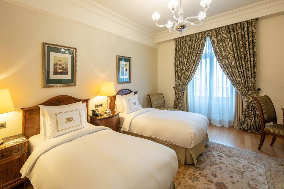 A hotel room with two beds, a chandelier, a large window with curtains, and a wooden nightstand with a lamp.