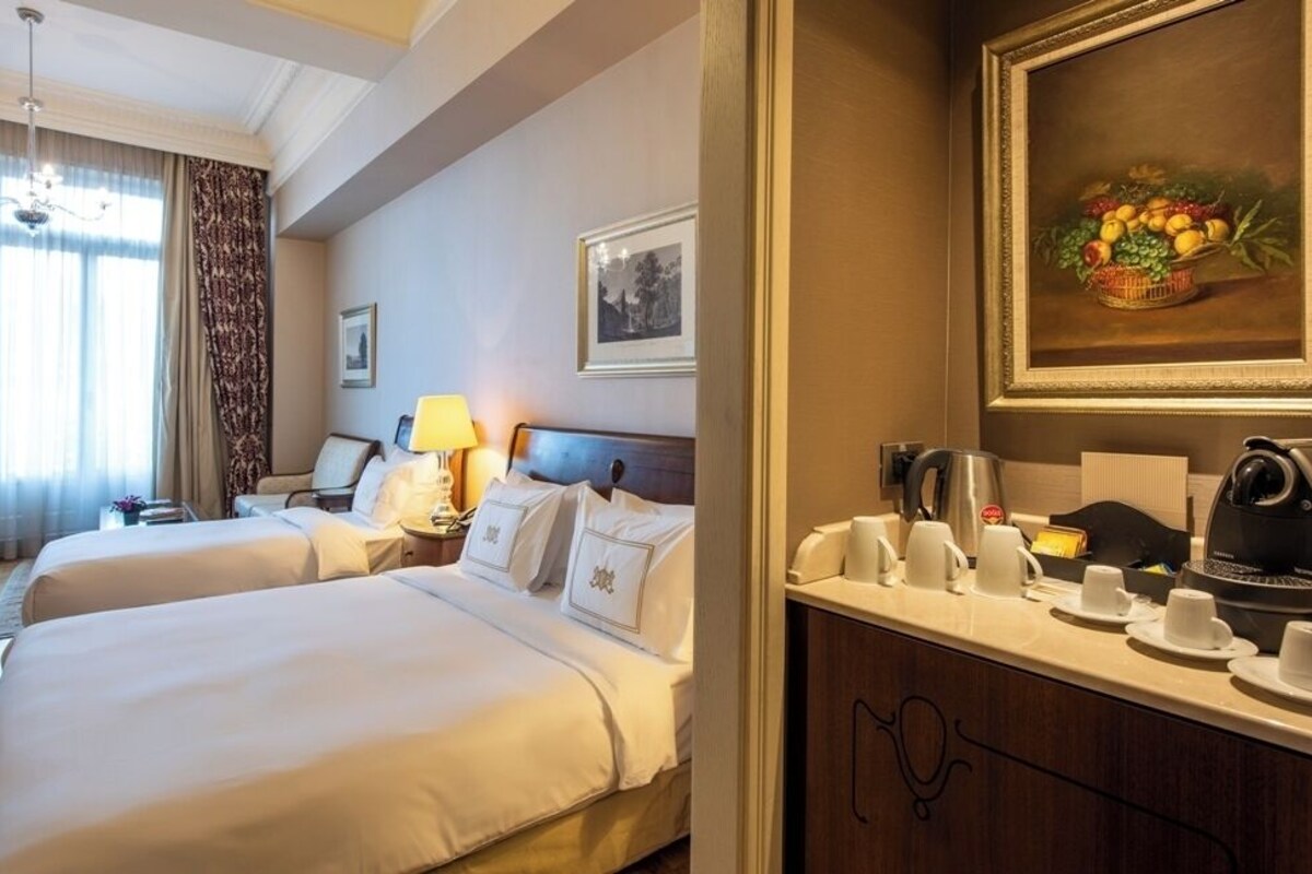 A hotel room with two beds, a desk, a lamp, a framed picture, a fruit still life, and a coffee station.
