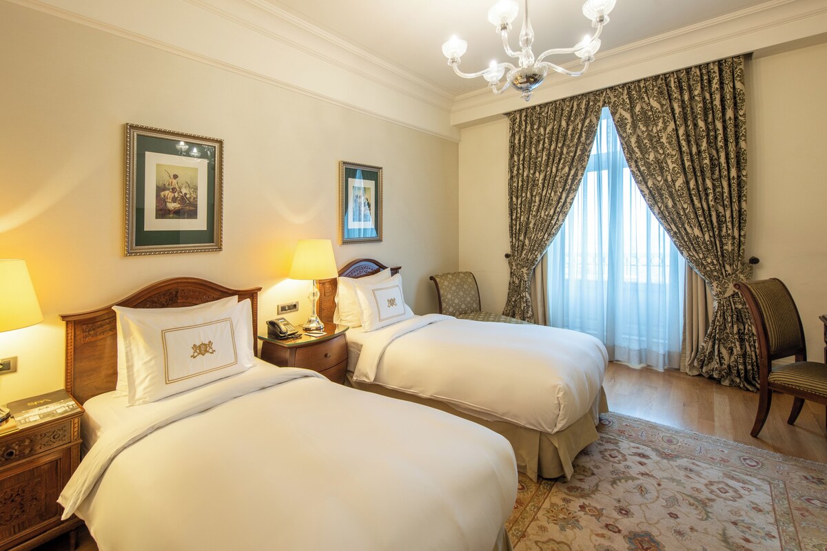 A hotel room with two beds, a chandelier, a large window with curtains, and a wooden nightstand with a lamp.