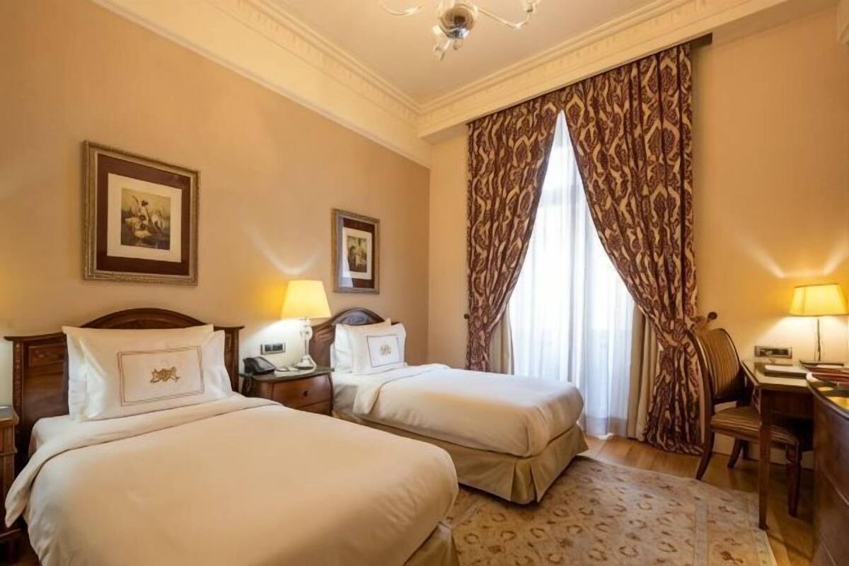 A hotel room with two beds, a desk, a chair, and a large window with curtains.