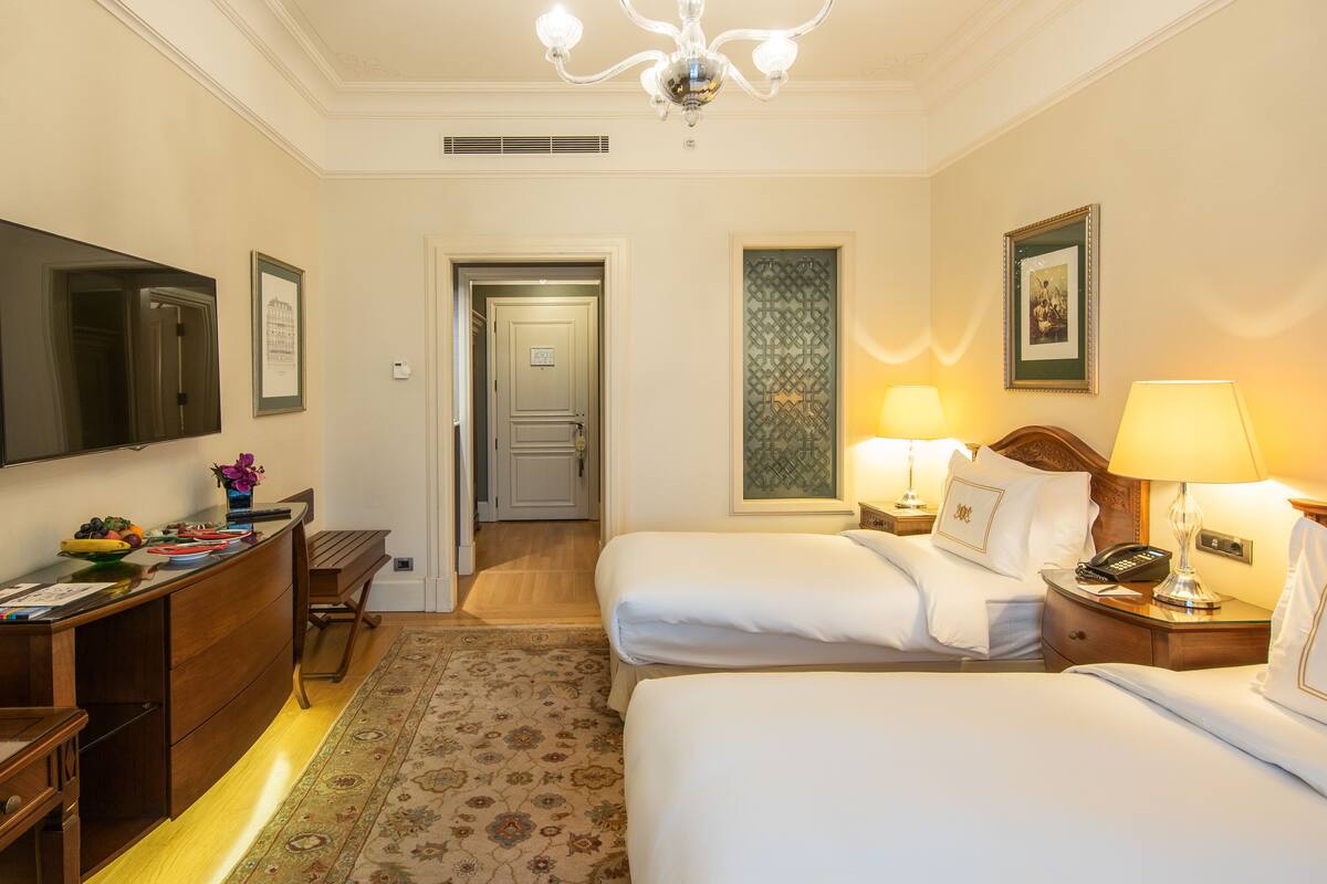 A hotel room with two beds, a desk, a TV, and a chandelier.