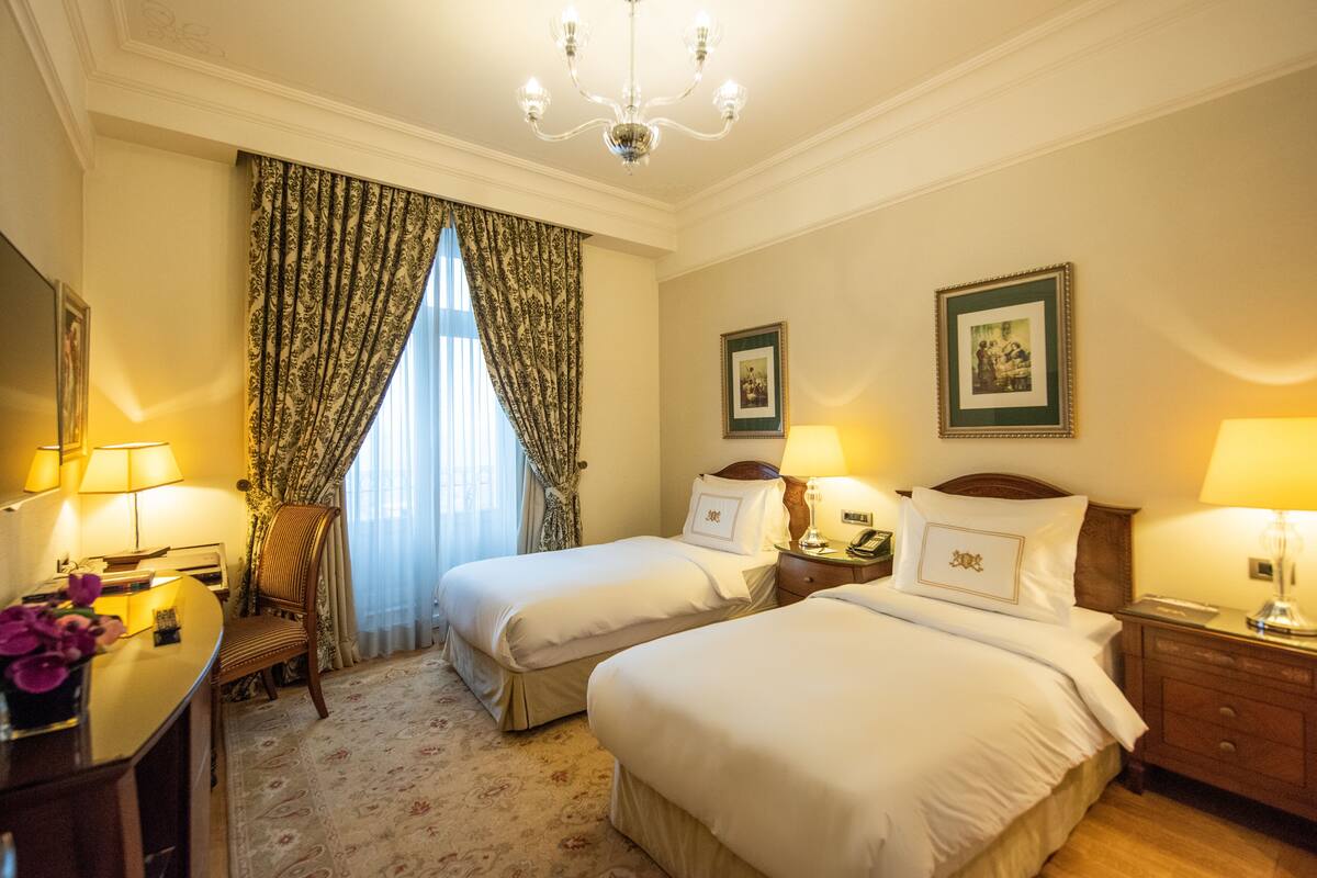 A hotel room with two beds, a desk with a lamp, a chair, and a desk lamp. There are two framed pictures on the wall.
