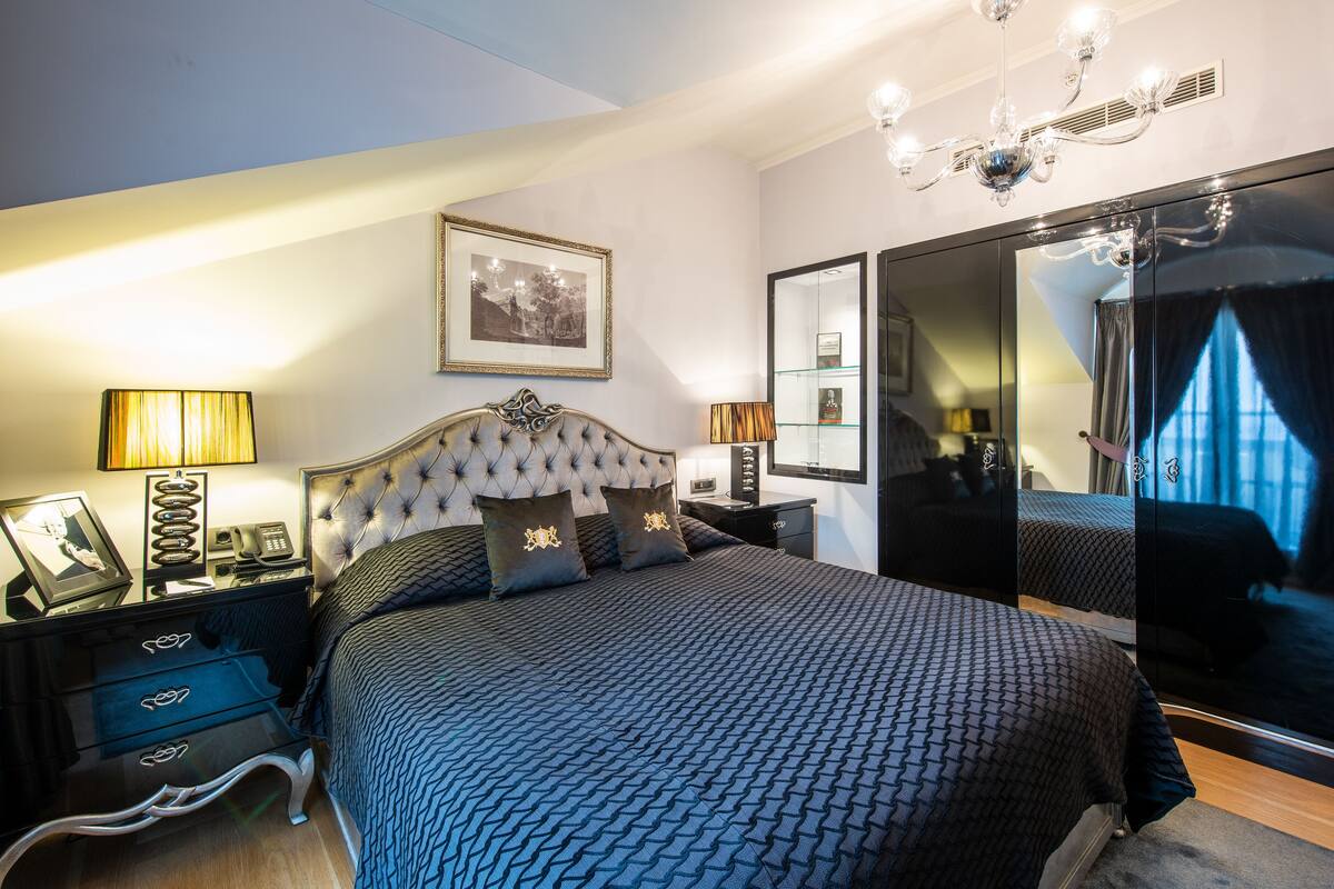 A bedroom with a large bed, two bedside tables, a dresser, a mirror, and a chandelier.