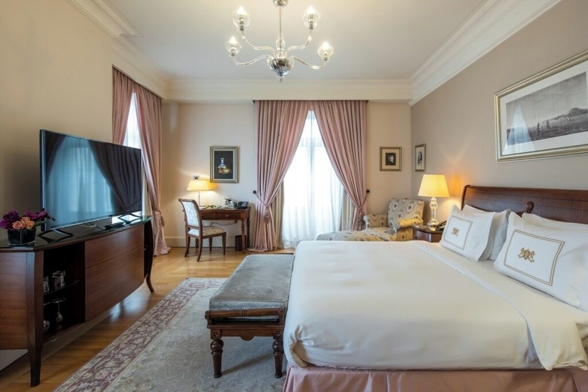A hotel room with a large bed, a desk with a chair, a television, and a chandelier.