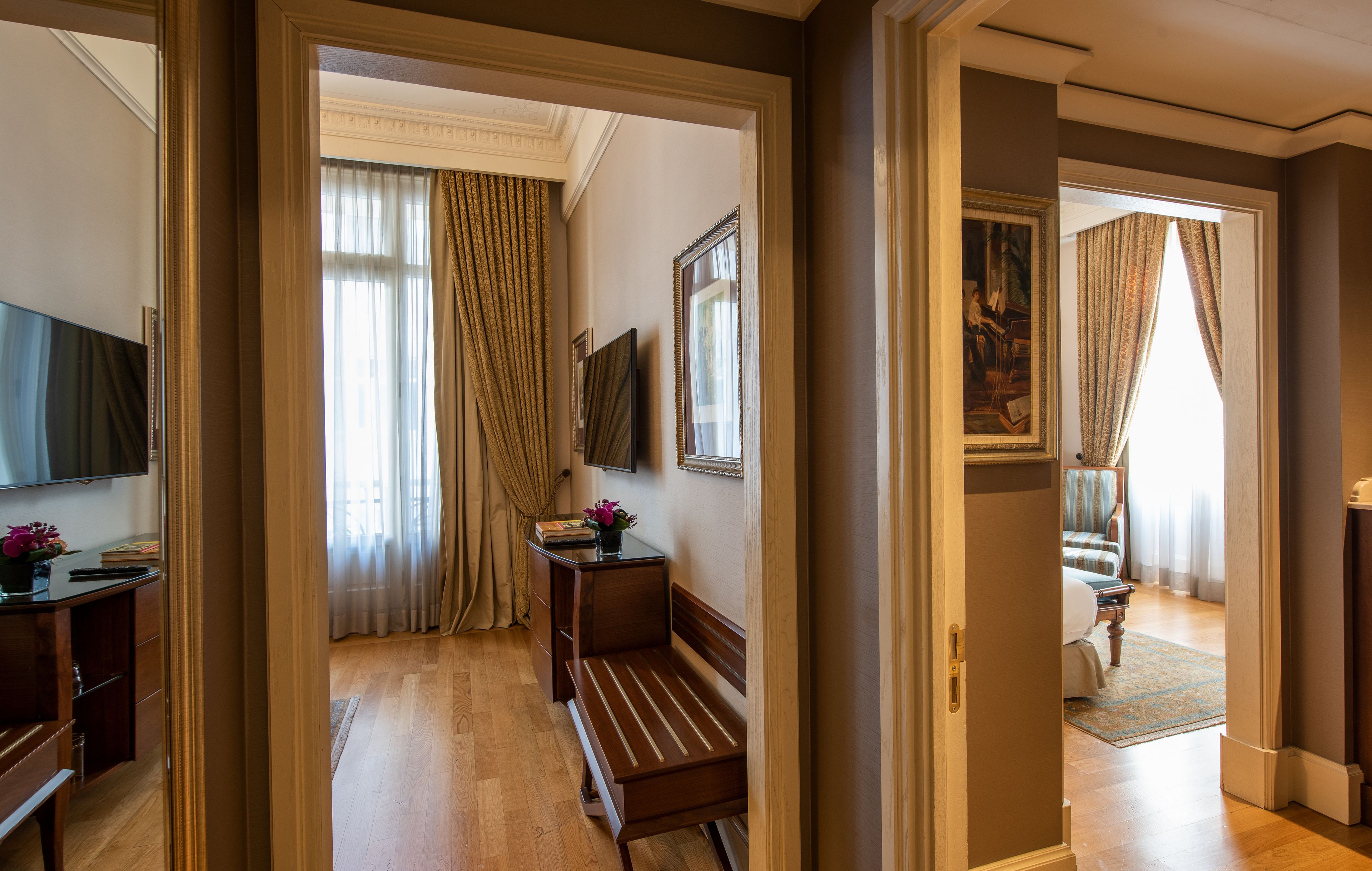 A hotel room with a wooden bench, a desk with a flat-screen TV, a painting on the wall, and a view of another room with a bed and curtains.
