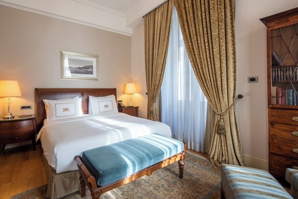 A hotel room with a large bed, two bedside tables with lamps, a wooden bench, a large window with curtains, and a wooden cabinet with drawers.