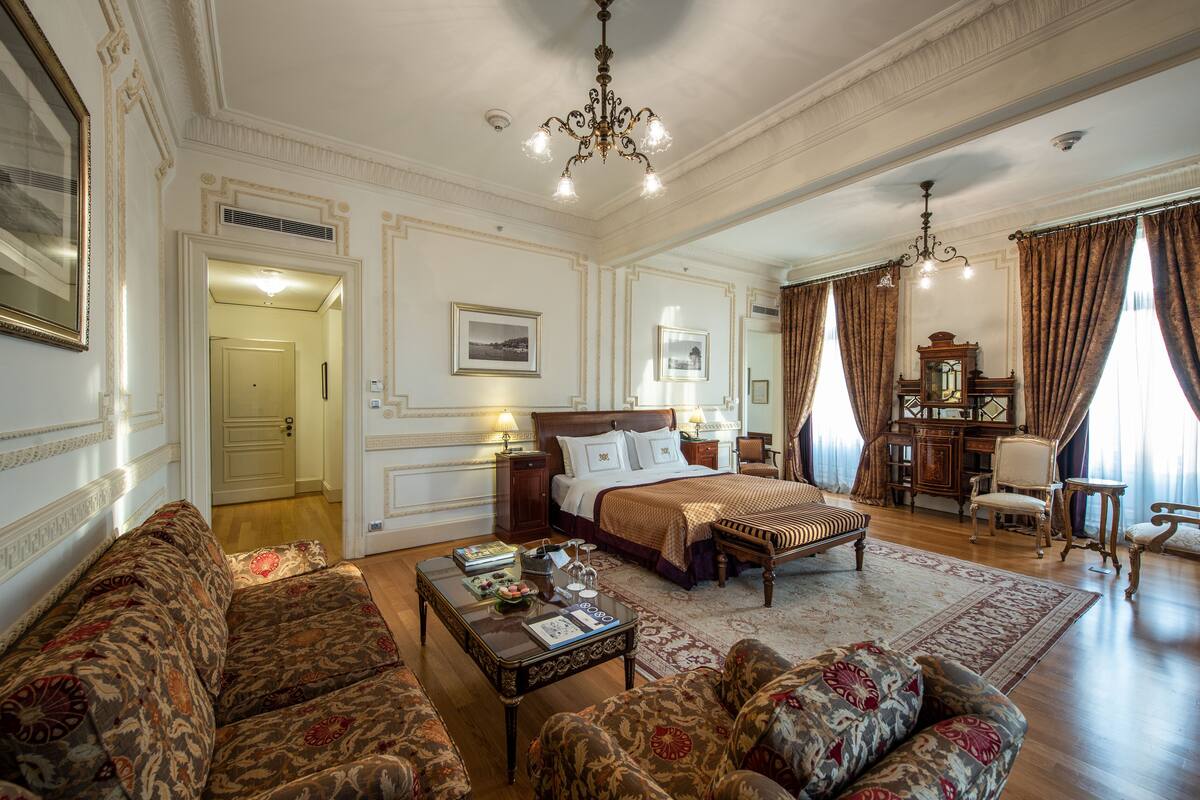 A spacious bedroom with a large bed, a sitting area with sofas, a wooden desk, and a chandelier.