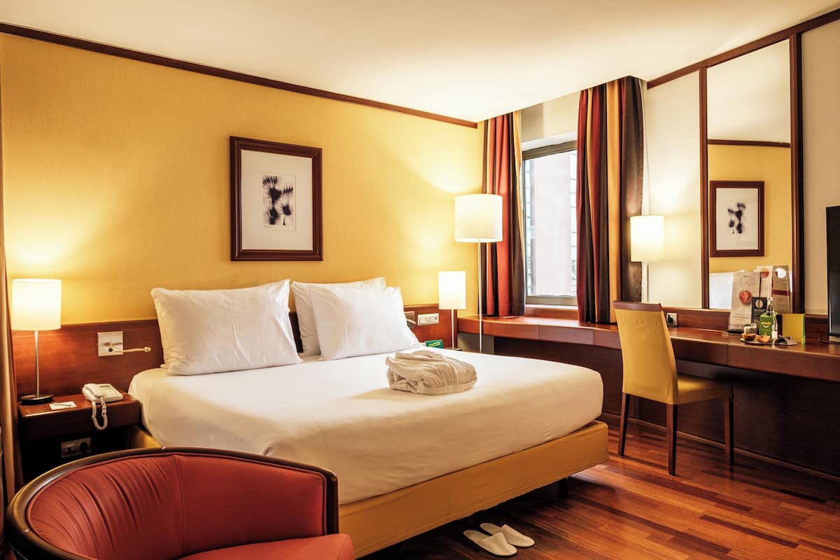 A hotel room with a large bed, a desk, a chair, a mirror, and a nightstand with a lamp.