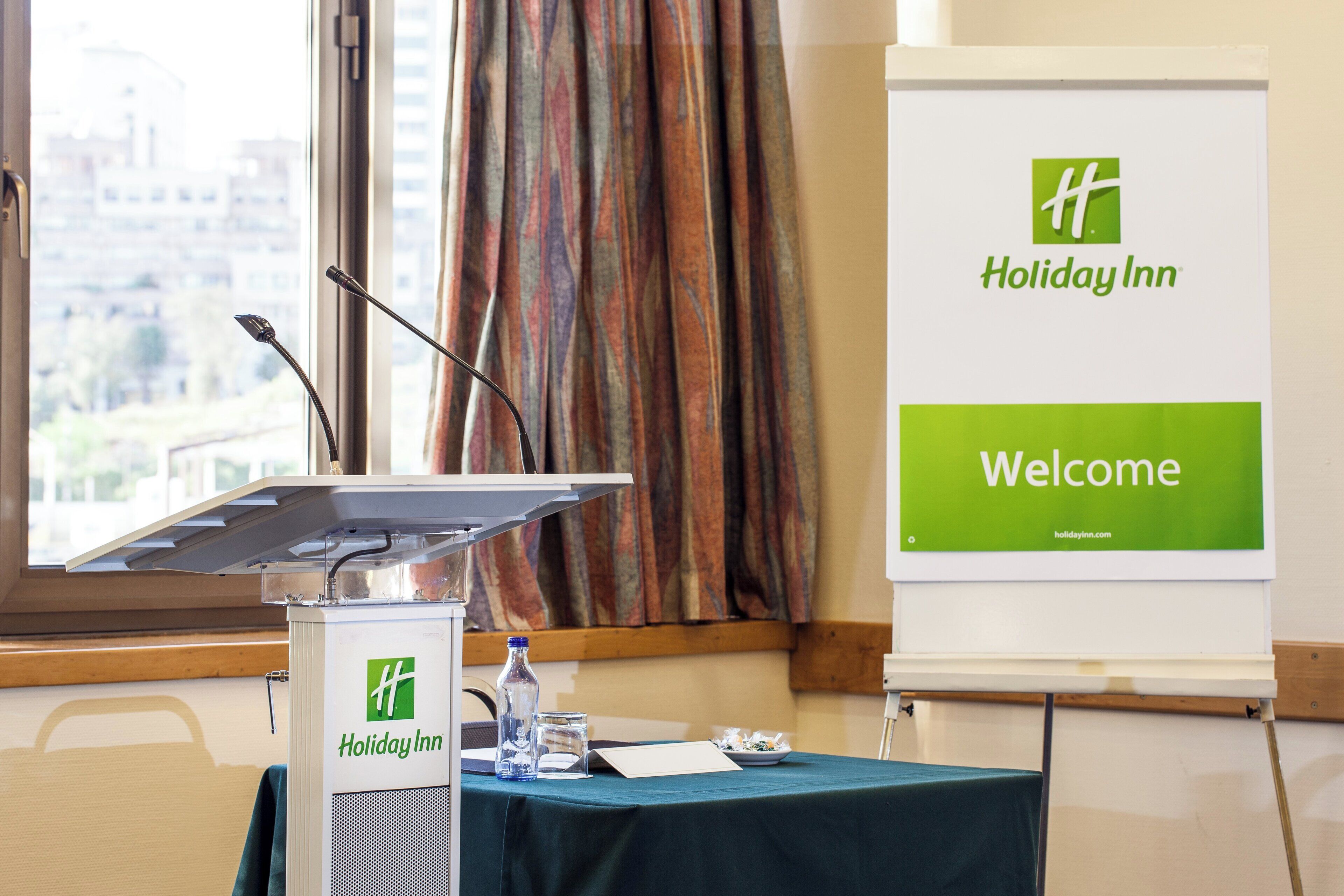 Een conferentiezaal van Holiday Inn met een spreekgestoelte, microfoon en een bord met de tekst 'Welcome'.