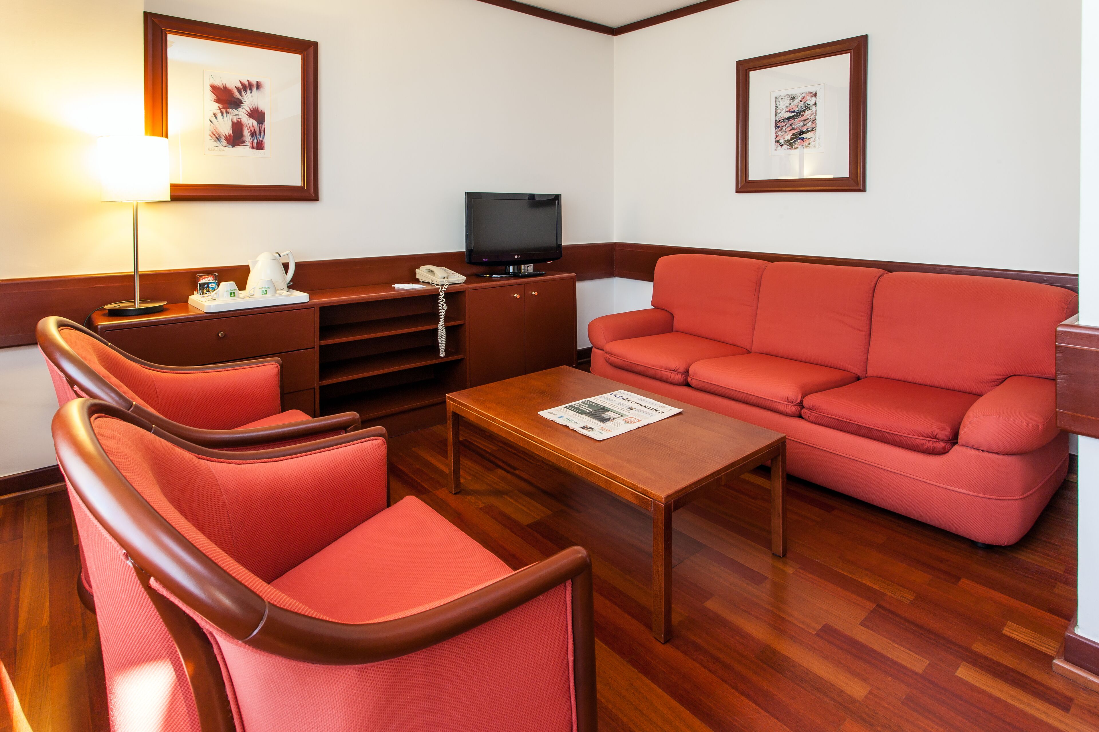 A hotel room with a red sofa, two armchairs, a wooden coffee table, a TV, and a cabinet with a telephone.