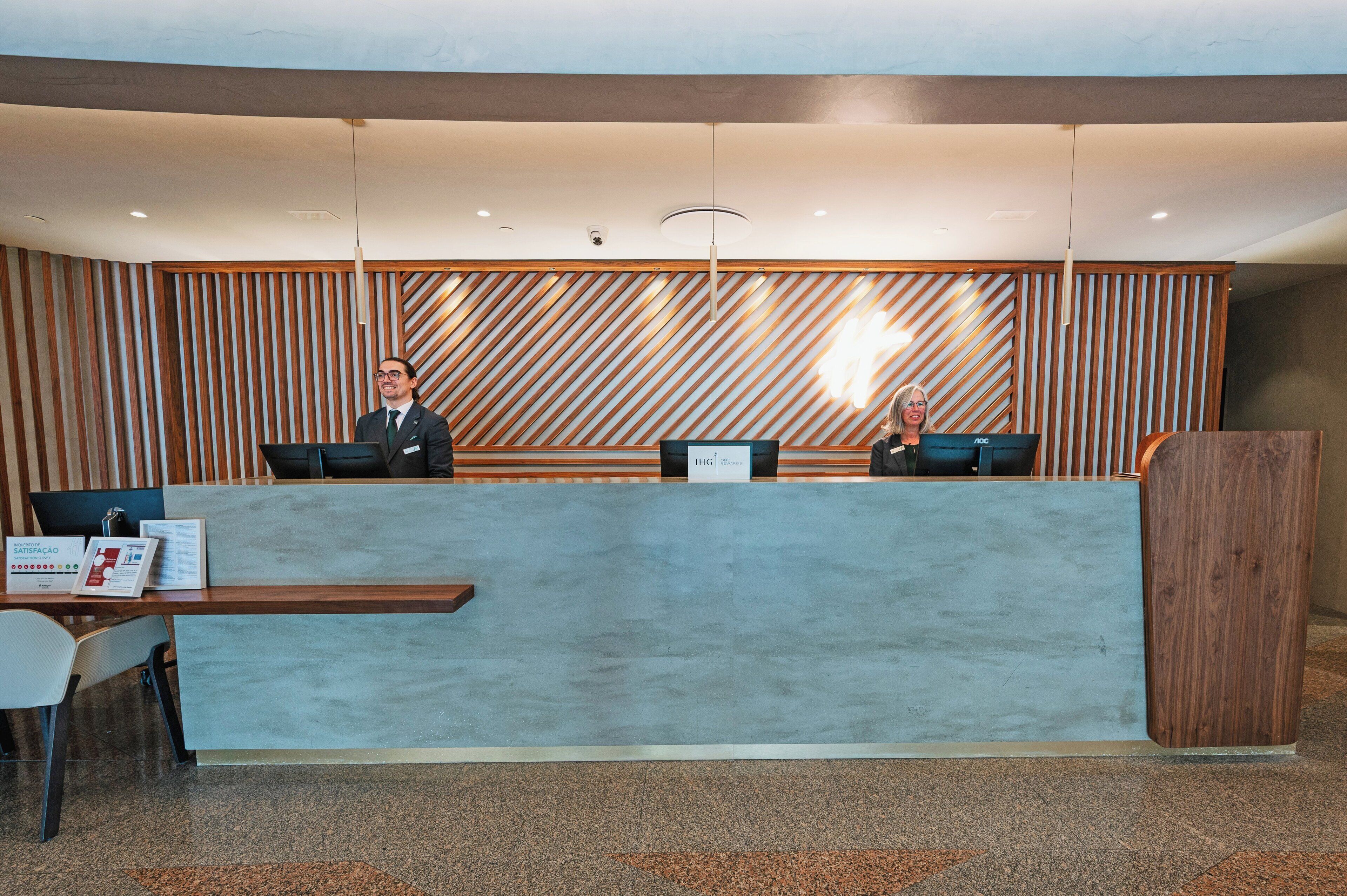 A reception desk with two staff members seated behind it, a wooden panel wall, and a modern light fixture.