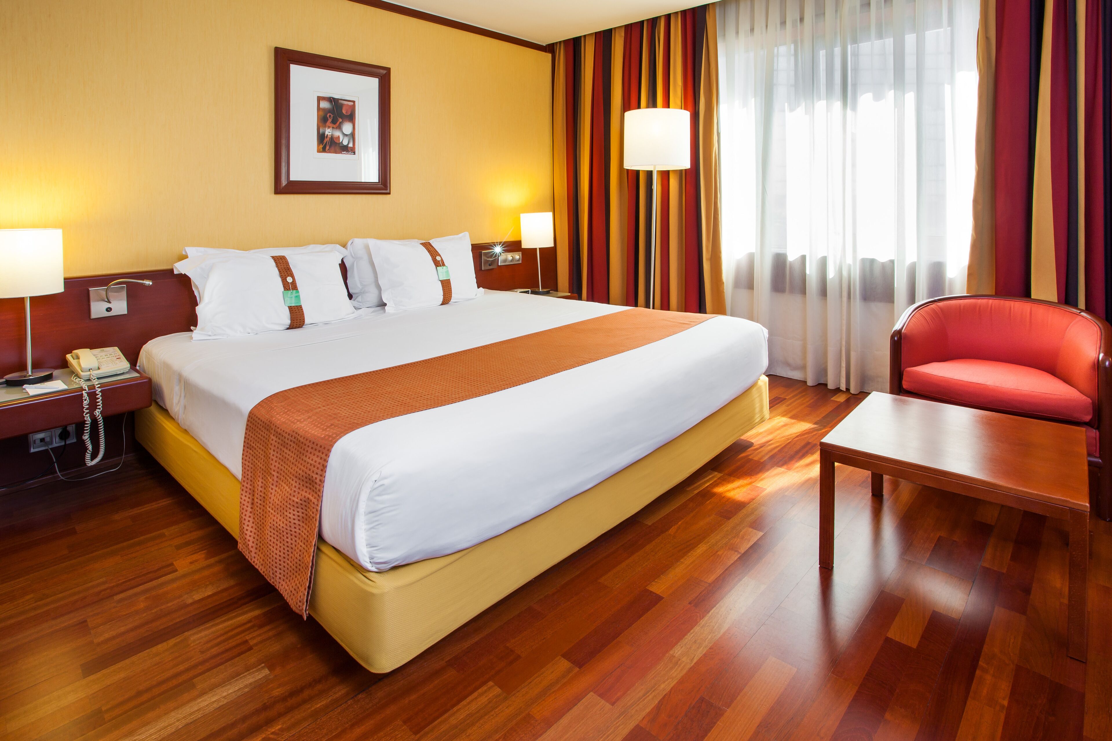 Habitación de hotel con una cama grande, lámparas de noche, un teléfono, una silla roja y un cuadro en la pared.