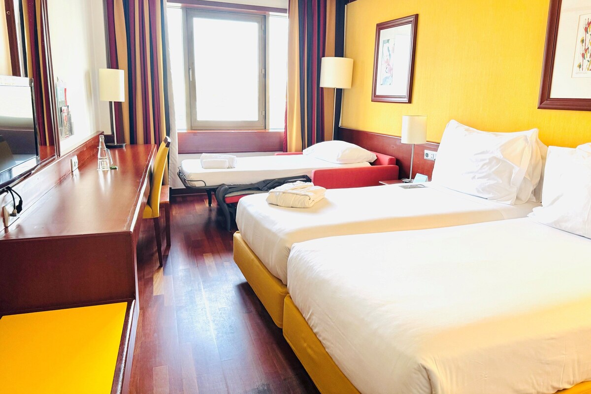 A hotel room with two beds, a desk, a chair, a window, and a lamp.