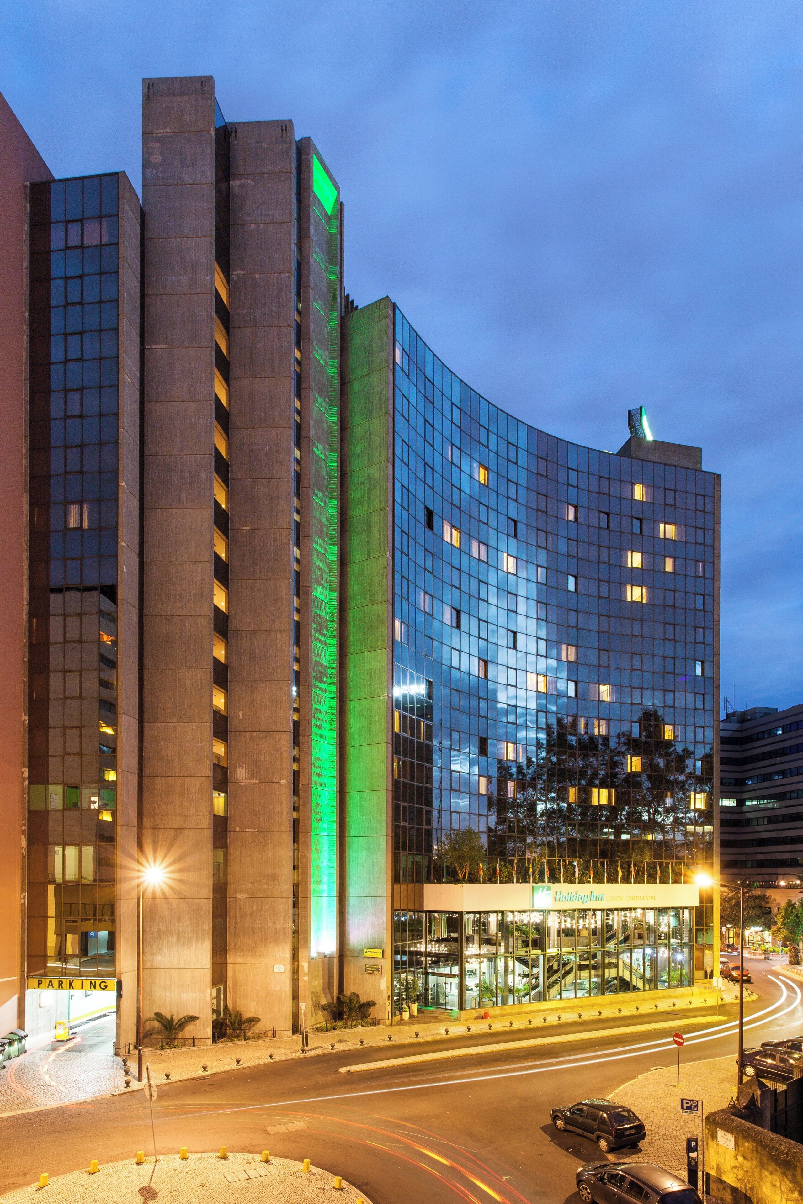 Et moderne hotel med en buet glasfacade, et markant, grønt oplyst lodret element og en parkeringsplads med afmærkede parkeringsbåse.