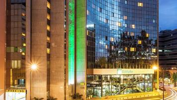 Et moderne hotel med en buet glasfacade, et markant, grønt oplyst lodret element og en parkeringsplads med afmærkede parkeringsbåse.