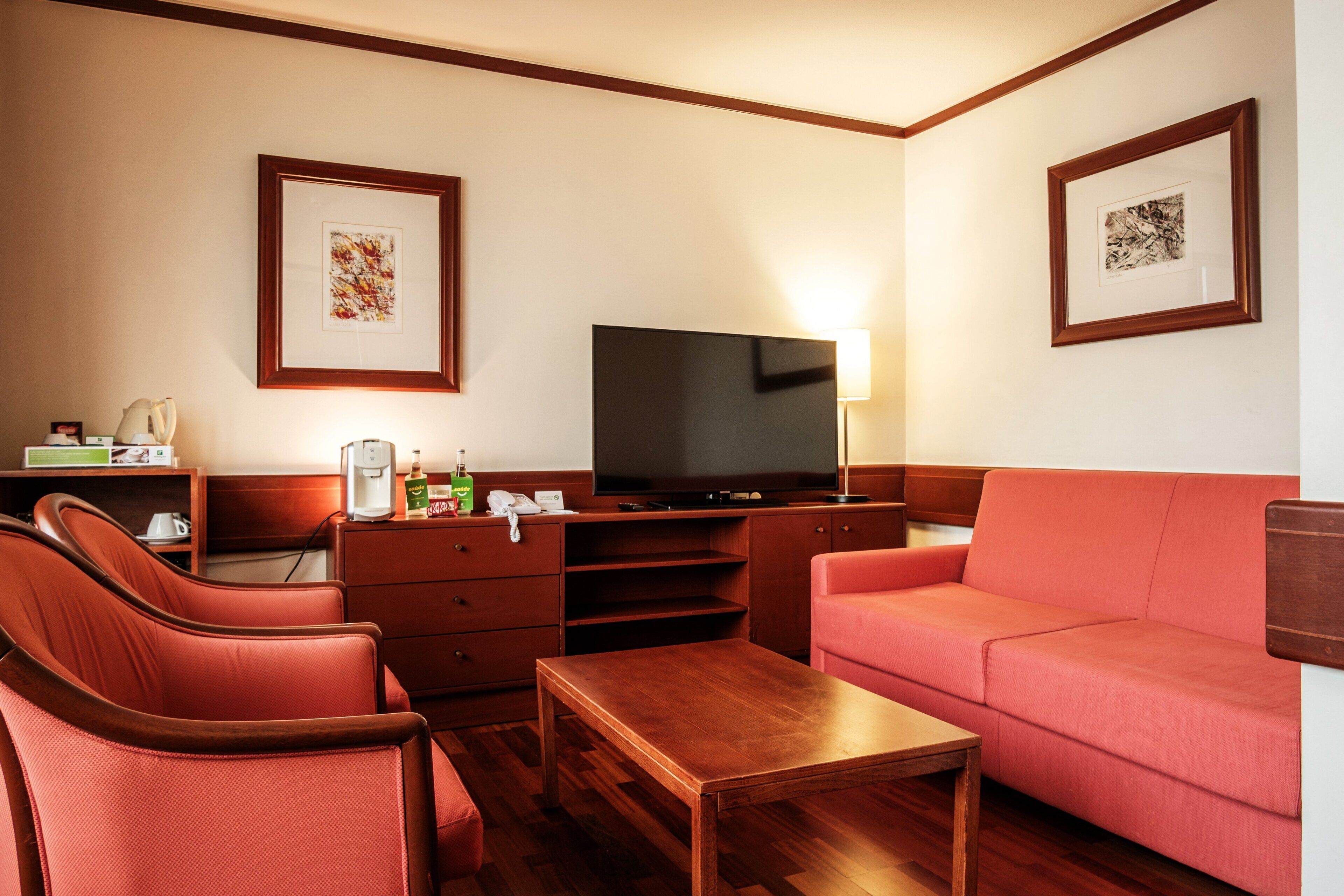 A hotel room with a television, a sofa, two armchairs, a wooden coffee table, a desk with a telephone, and framed artwork on the walls.