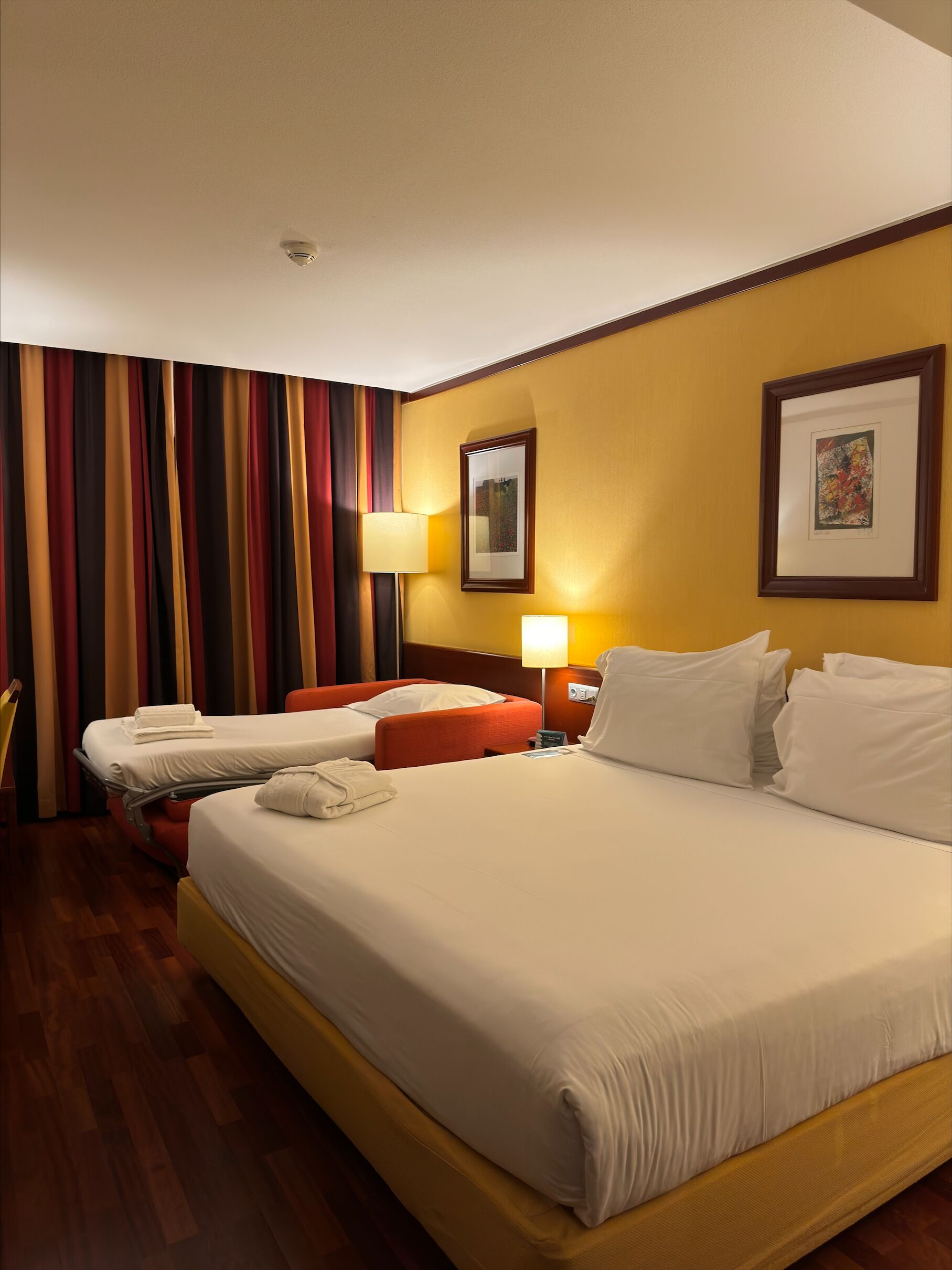 A hotel room with two beds, a wooden floor, a striped curtain, and two wall-mounted lamps.