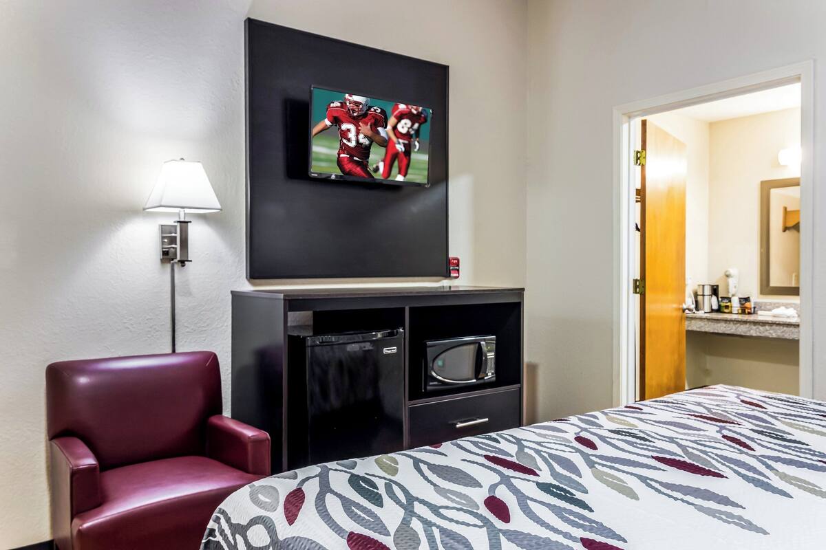 Habitación de hotel con cama, televisor montado en la pared, escritorio con microondas y una silla roja.
