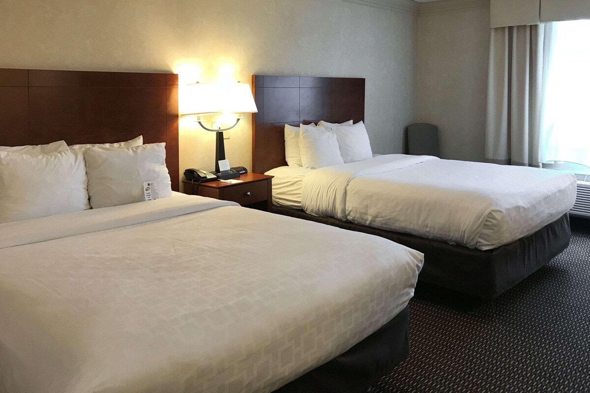 A hotel room with two beds, a nightstand with a lamp, and a window with curtains.