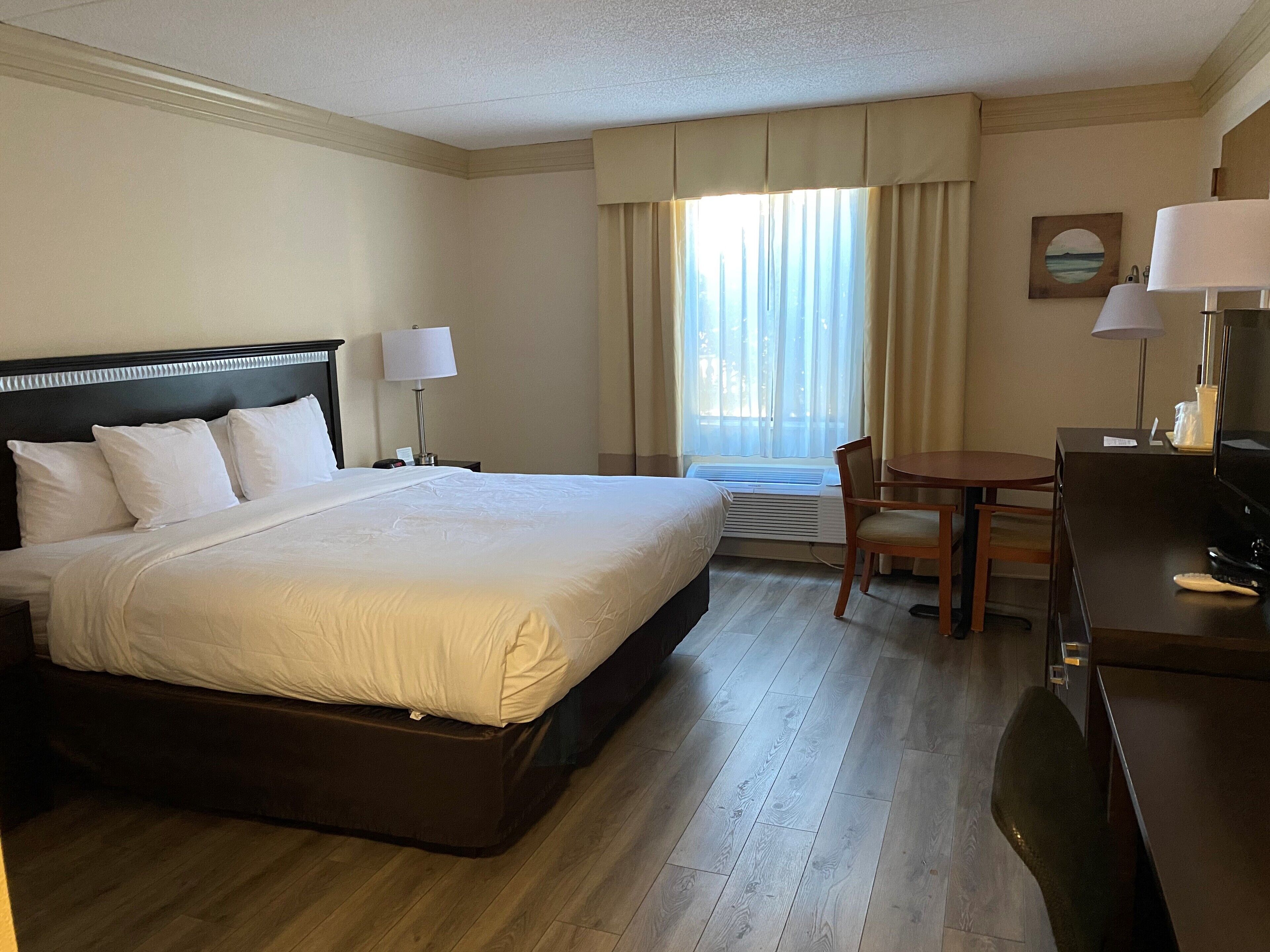 A hotel room with a large bed, a desk, a chair, a lamp, a picture on the wall, and a window with curtains.