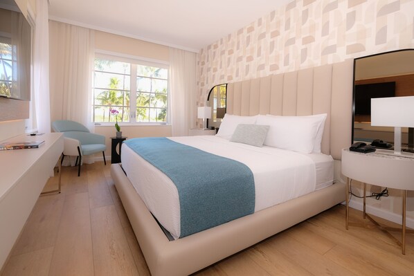 A modern hotel room with a large bed, a desk, a chair, a bedside table with a lamp, and a large window with a view of palm trees. - Avalon Hotel (Miami Beach)