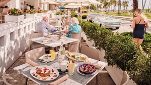 An outdoor dining area with tables, chairs, umbrellas, and a view of palm trees and a street. - Avalon Hotel (Miami Beach)